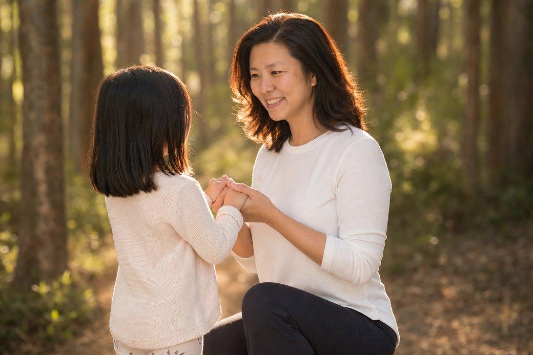 Dao, the founder of PlotJoys, kneeling in sunlit woodland holding both of her young daughter’s hands
