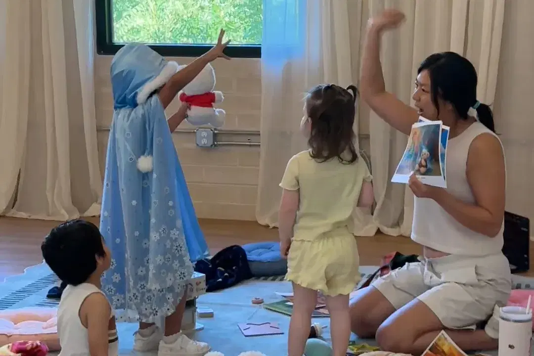 Story time mid-spell — a girl in a snow-queen cape raises her bear while Dao tells the story, printed pages in hand